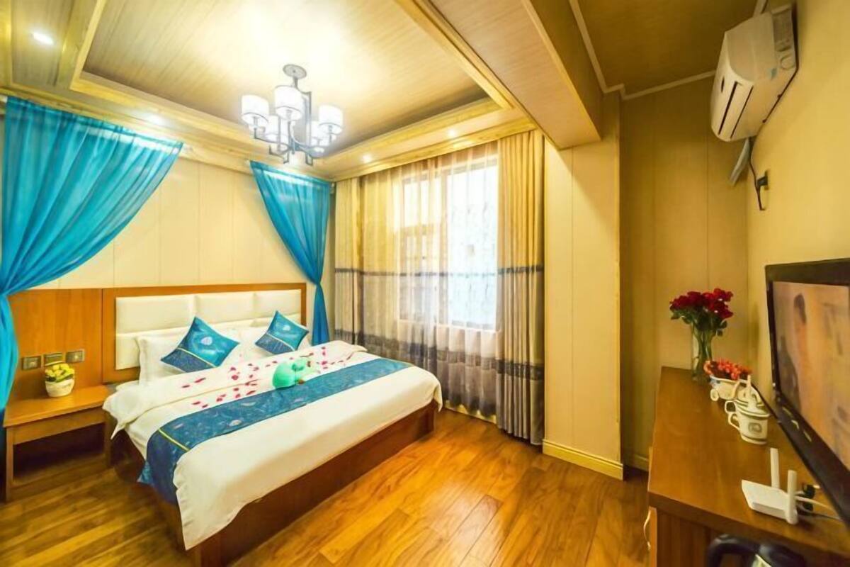 A hotel room with a wooden floor, a bed with blue and white bedding, a nightstand with a lamp, a TV, and a wall-mounted air conditioner.