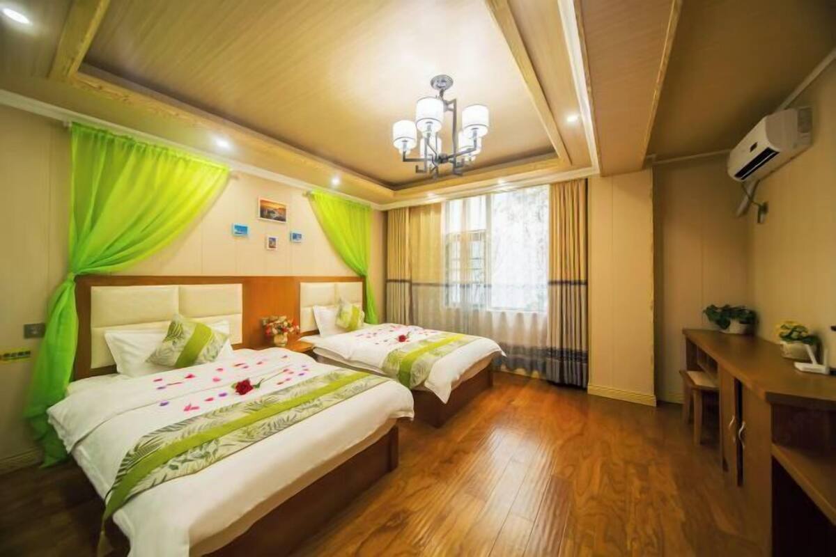 A hotel room with two beds, wooden flooring, a chandelier, a desk, and a window with curtains.