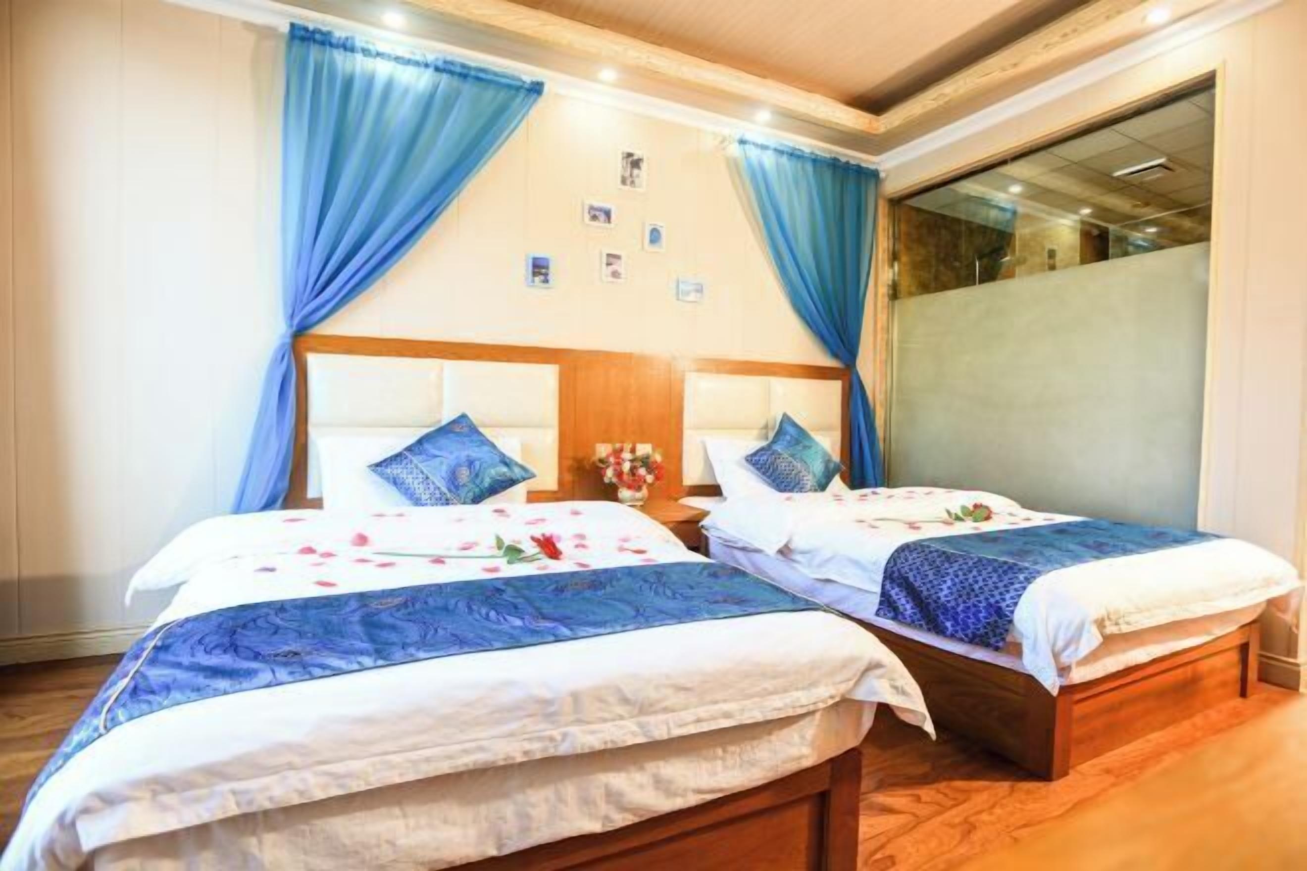 Two single beds with headboards, a nightstand with a vase, and a window with blue curtains.