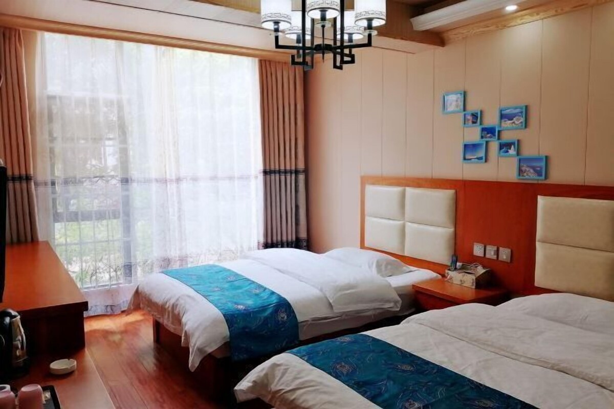 A hotel room with two beds, a wooden desk, a chandelier, and a large window with curtains.