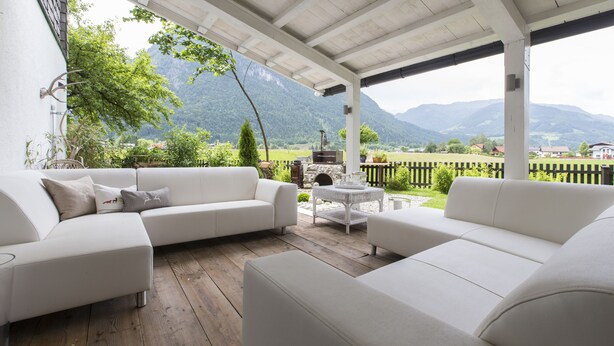 Ein überdachter Außenbereich mit weißen Sofas und einem kleinen runden Tisch, mit Blick auf eine gebirgige Landschaft.