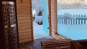Eine Holzhütte mit großem Fenster, Kamin aus Stein und Blick auf eine verschneite Berglandschaft.