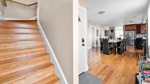 Una escalera de madera que lleva a una habitación con una puerta, un comedor con mesa y sillas, y una cocina con electrodomésticos.