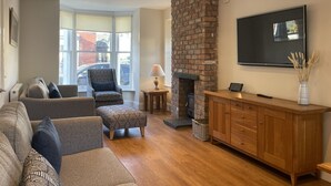 A living room with a fireplace, a TV mounted on the wall, and a large window providing a view of the street.