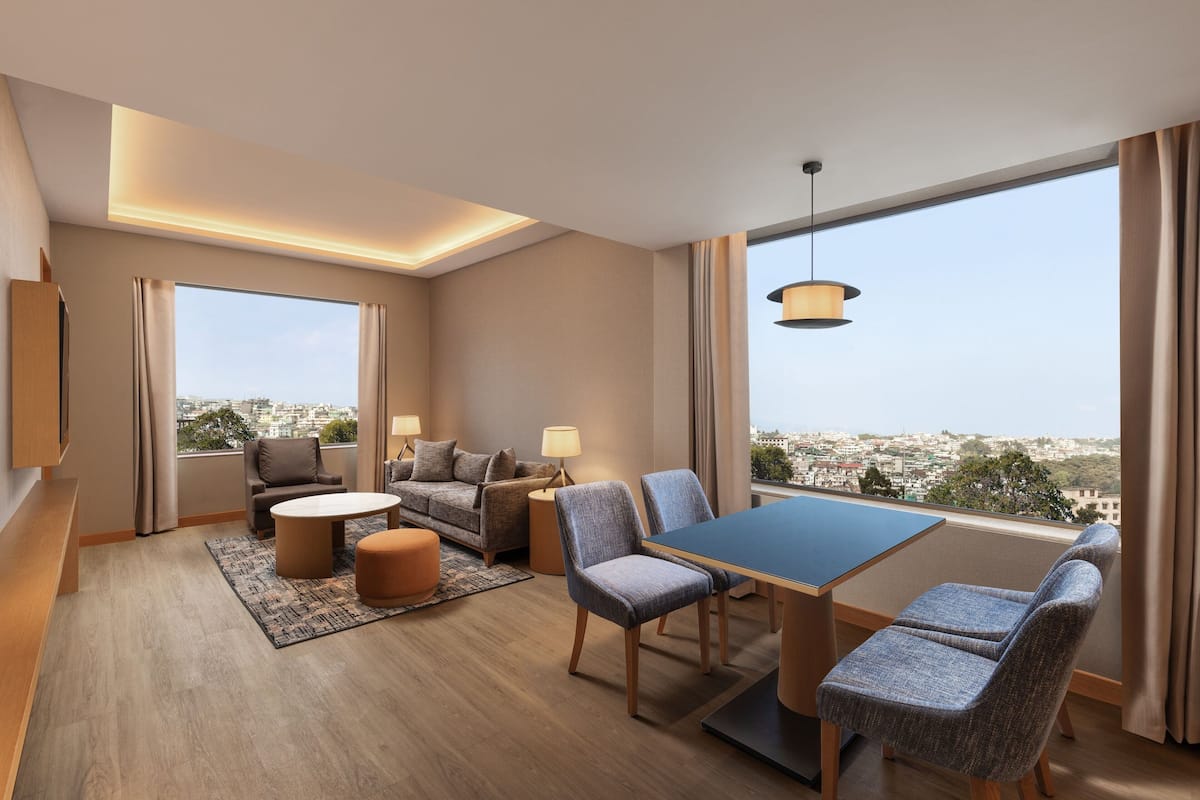 A modern hotel room with a large window, a sofa, a dining table, and a view of the city.