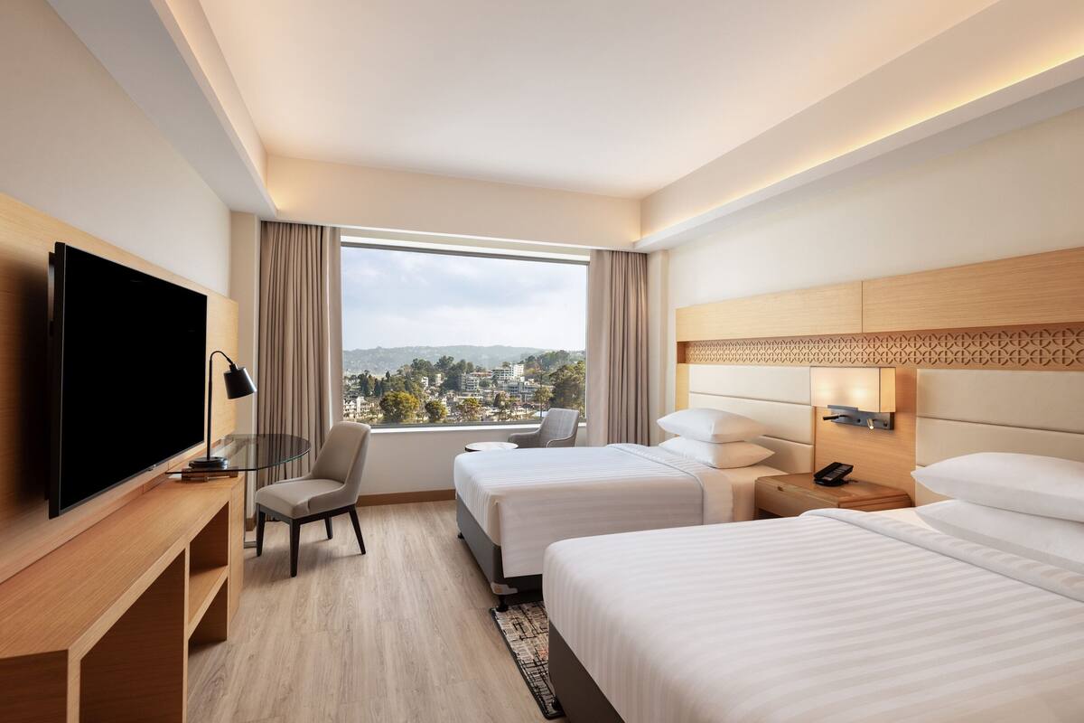 A hotel room with two beds, a TV, a desk, and a view of a cityscape.