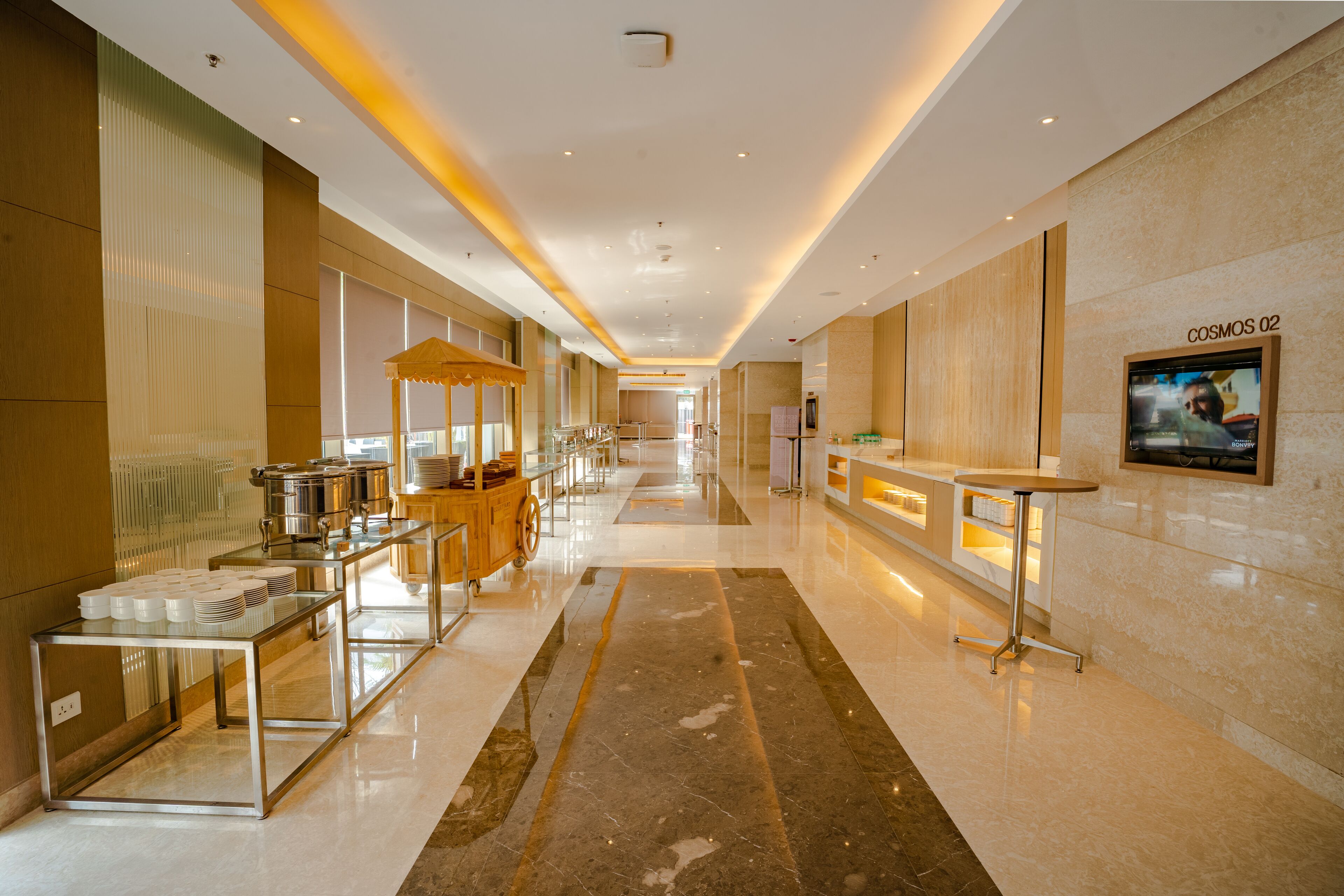 Een moderne hotelgang met een buffetgedeelte, een receptiebalie en een televisiescherm waarop een persoon te zien is.