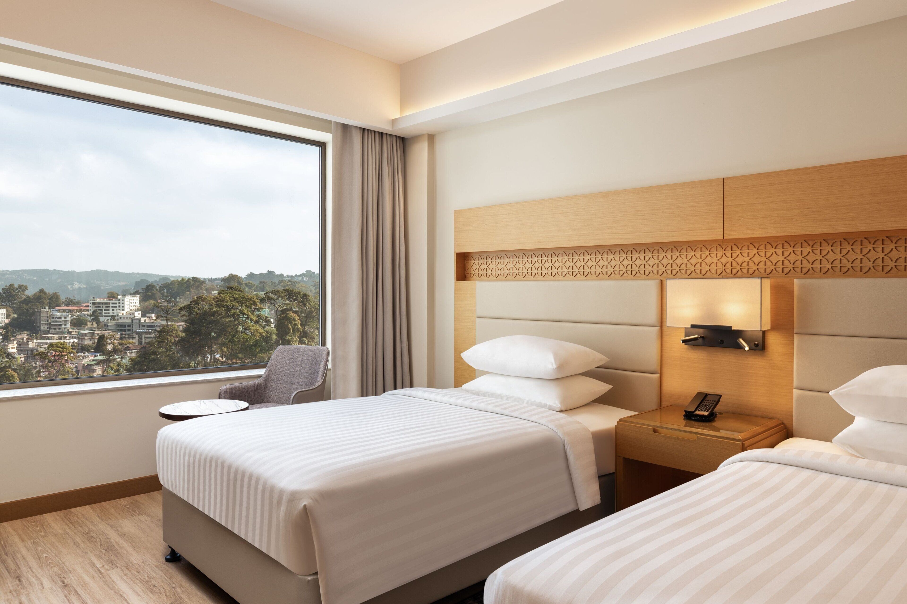 Ein Hotelzimmer mit zwei Betten, einem Nachttisch mit Telefon, einem Sessel, einem kleinen Tisch und einem großen Fenster mit Stadtblick.