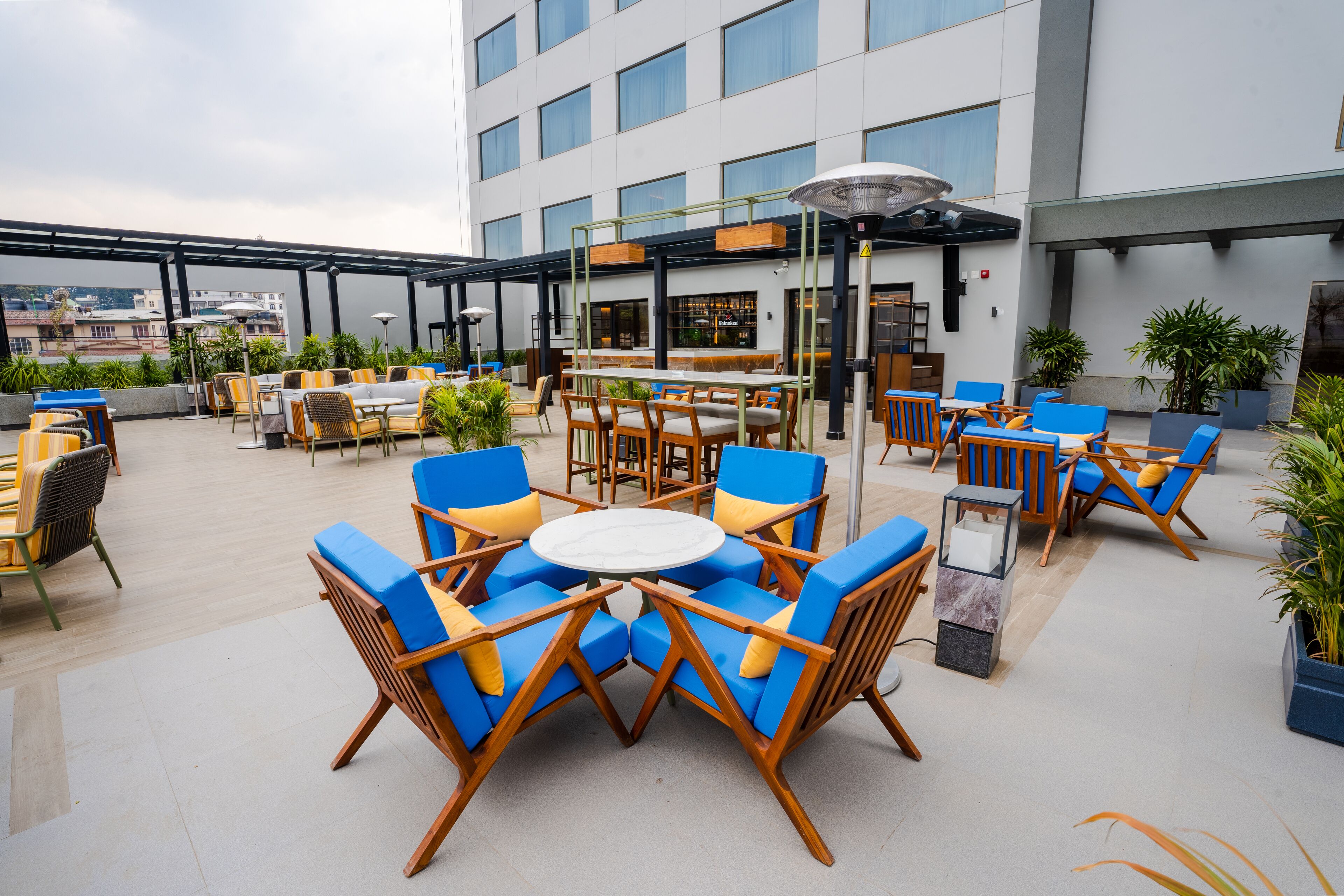 Un patio en la azotea con mesas y sillas al aire libre, incluyendo sillas azules y amarillas, y una zona de bar con vistas a la ciudad.