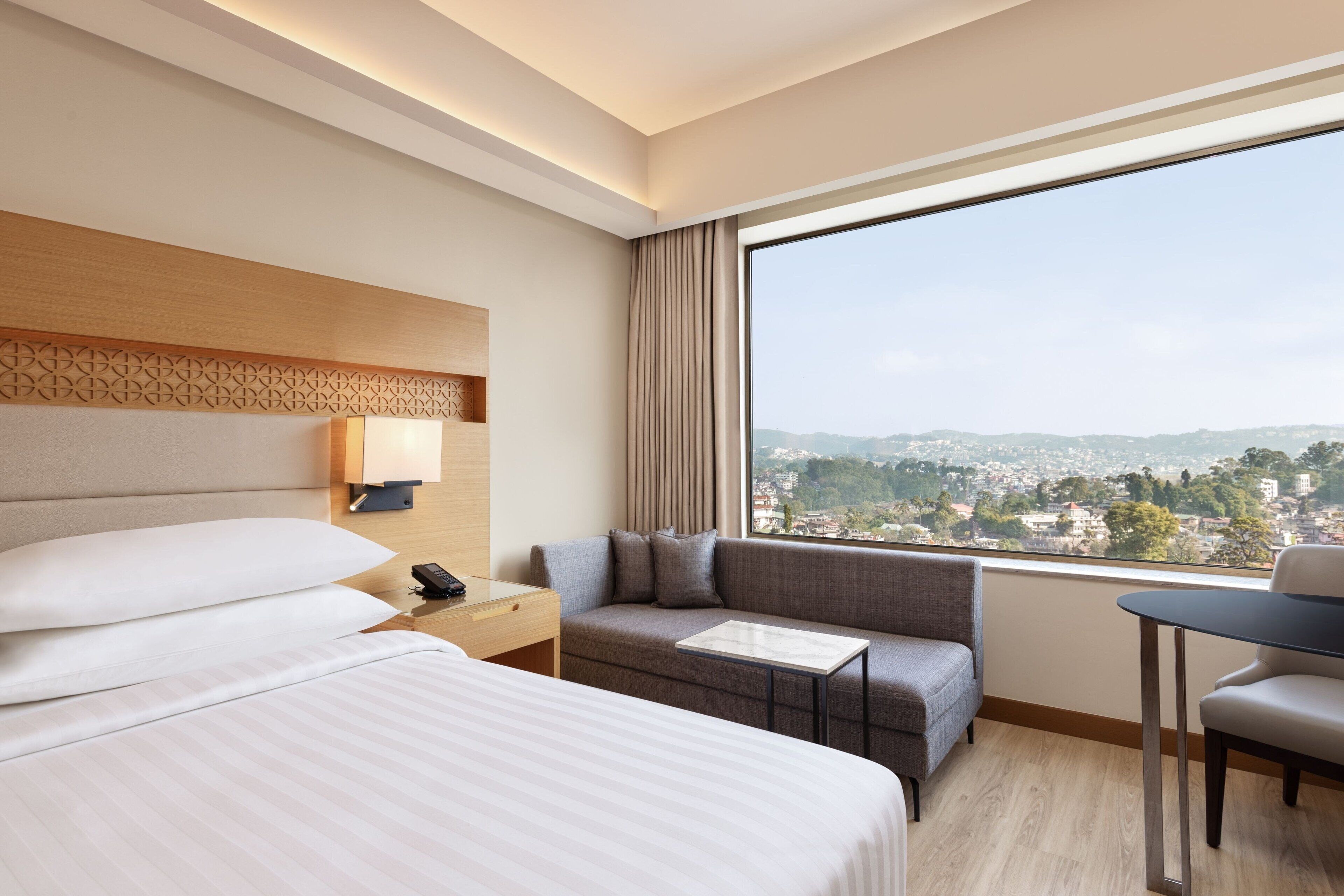 A hotel room with a large bed, a sofa, a small table, and a view of a cityscape.