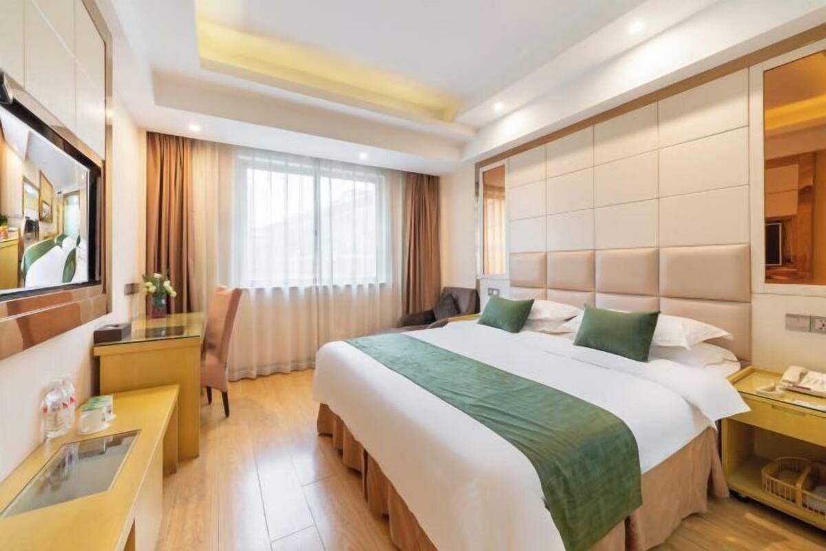 Une chambre d’hôtel avec un grand lit, un bureau avec une chaise, une table de chevet, un miroir et une fenêtre avec des rideaux.