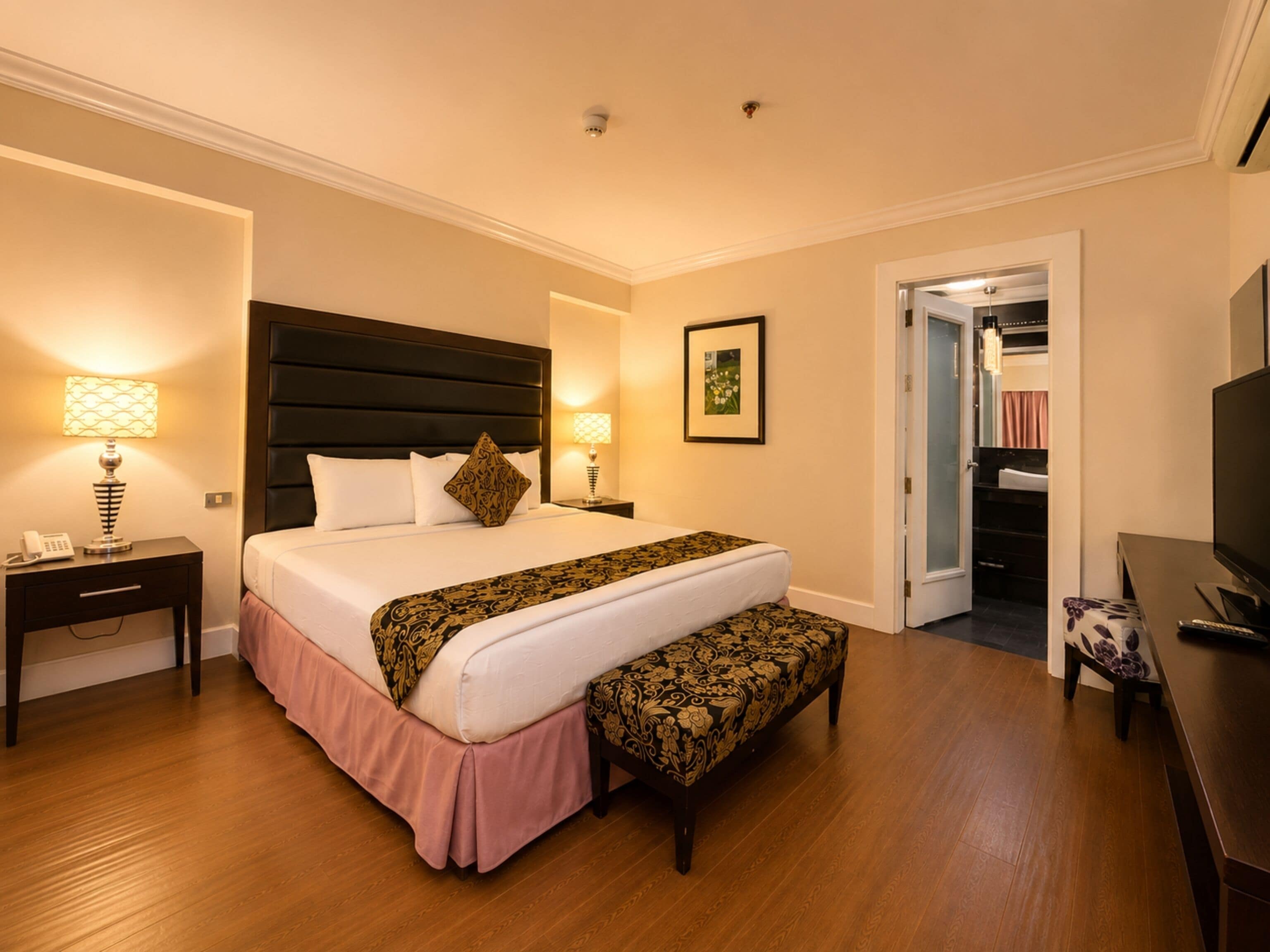 Habitación de hotel con una cama grande, lámparas de noche, un banco, un televisor y una puerta que da a un armario.