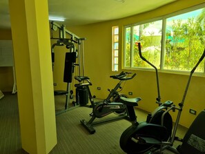 A gym with exercise equipment, a window with a view of greenery, and a yellow wall. - Fiesta Garden Hotel (Bantay)