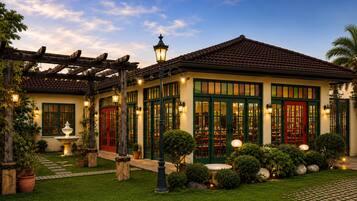 Un jardín cuidado con una fuente, una pérgola y un edificio con tejado de baldosas y ventanas con marco verde.