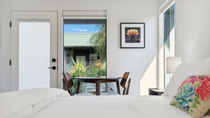 A bedroom with a bed, a dining table with chairs, a potted plant, a framed picture, and a window with a view of greenery. - SPCH07 - Eco-friendly Studio Container - 80 Walkscore! (Phoenix)