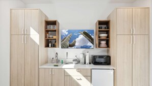 A modern kitchen with light wood cabinets, a microwave, and a sink. There are two wooden shelves above the sink, one holding cups and the other holding plates. A large window above the sink allows natural light to enter. - SPCH07 - Eco-friendly Studio Container - 80 Walkscore! (Phoenix)