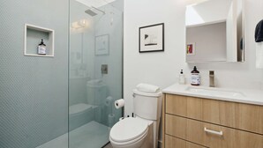 A modern bathroom with a glass shower enclosure, a toilet, a sink with toiletries, and a wooden cabinet. - SPCH07 - Eco-friendly Studio Container - 80 Walkscore! (Phoenix)