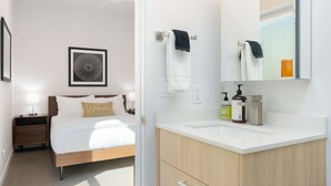 Ein modernes Hotelzimmer mit Bett, Nachttischen, einem gerahmten Kunstwerk, einem weißen Badezimmer mit Waschbecken, Pflegeprodukten und einem Spiegel.