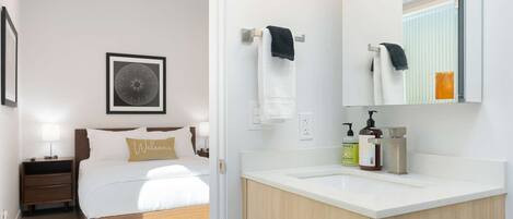 Une chambre d’hôtel moderne comprenant un lit, des tables de chevet, une œuvre d’art encadrée, une salle de bain blanche avec un lavabo, des articles de toilette et un miroir.