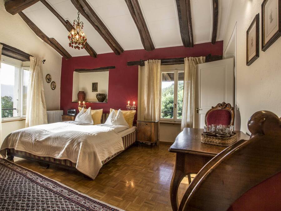 Une chambre à coucher avec un lit en bois, un mur rouge, un lustre et une vue sur la verdure à l’extérieur.