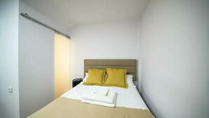 A small, neatly made bed with a beige headboard and two yellow pillows, a white blanket, and a folded white towel on the bed.