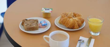 Una mesa con alimentos para el desayuno, incluyendo cruasanes, pan, una taza de café, un vaso de jugo de naranja y un pequeño recipiente de yogur.