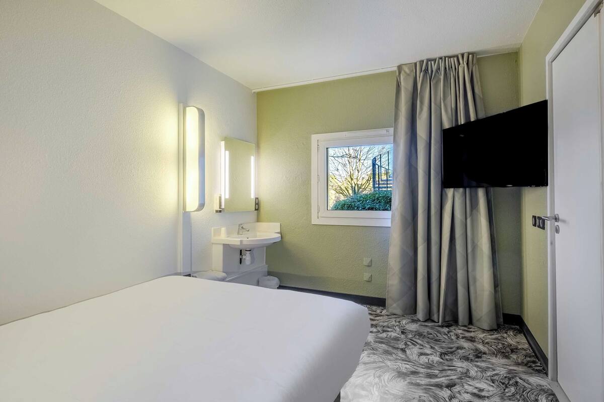 Ein Hotelzimmer mit Bett, Waschbecken, Spiegel, einem Fenster mit Vorhängen und einem an der Wand befestigten Fernseher.