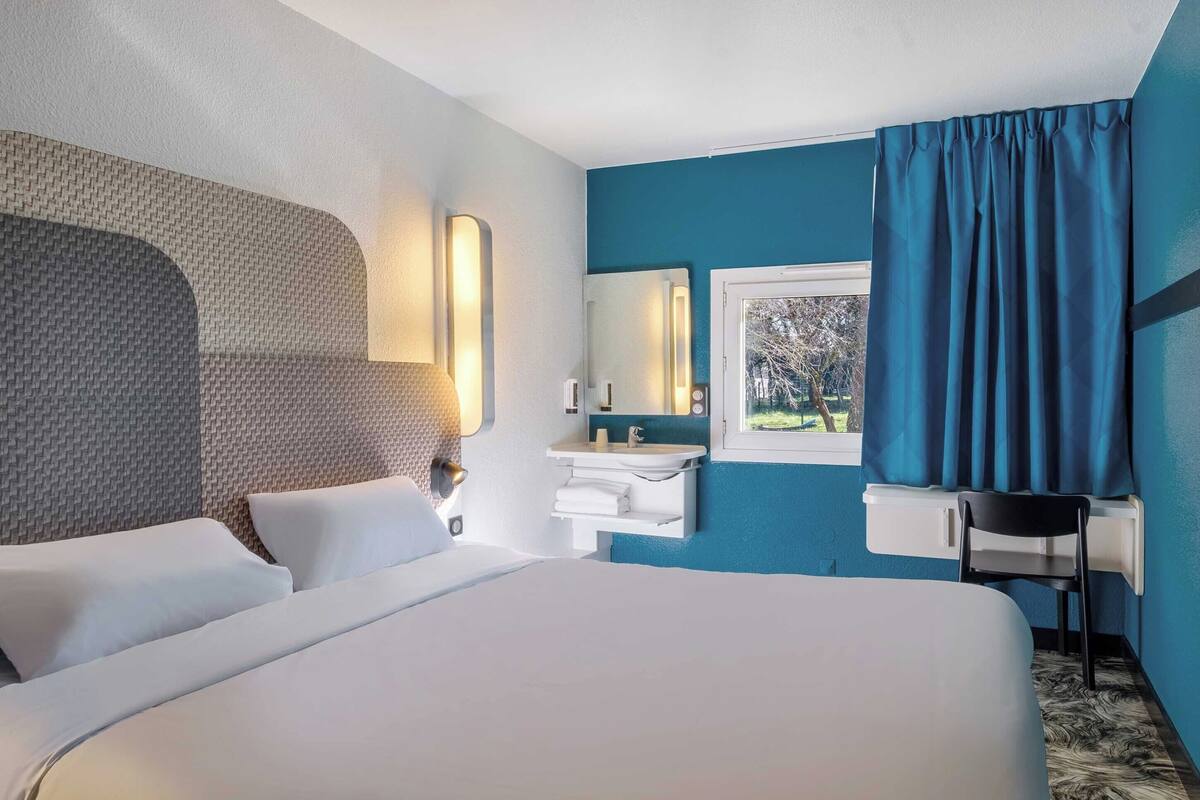 Ein Hotelzimmer mit einem großen Bett, einem Nachttisch, einem Spiegel, einem Fenster mit Vorhängen und Blick auf Bäume.