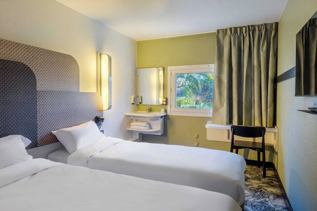 Ein Hotelzimmer mit zwei Betten, einem Fenster mit Vorhängen, einem Waschbecken und einem Sessel.