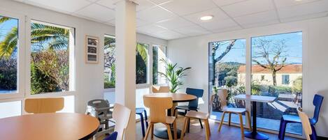 A modern indoor space with multiple tables and chairs, large windows with views of greenery and buildings, and a ceiling with recessed lighting.