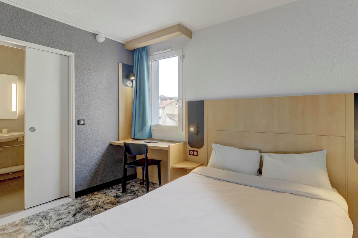 A hotel room with a bed, a desk, a chair, a mirror, and a window with curtains.