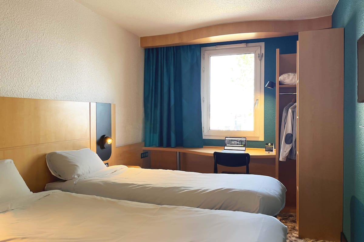 A hotel room with two beds, a desk, a chair, a wardrobe, and a window with curtains.