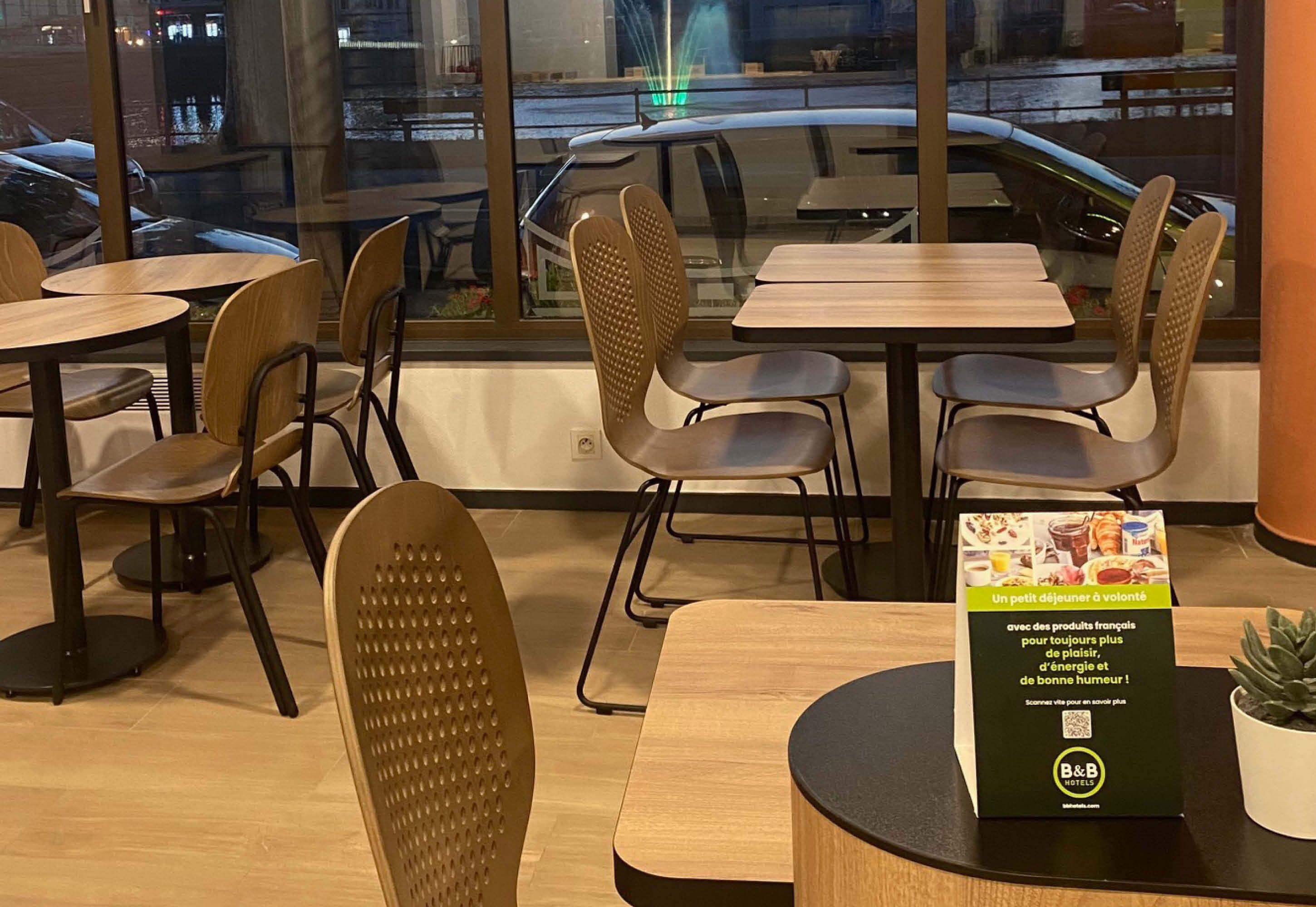 Een restaurant met houten tafels en stoelen, een menukaartstandaard en uitzicht op geparkeerde auto's door grote ramen.