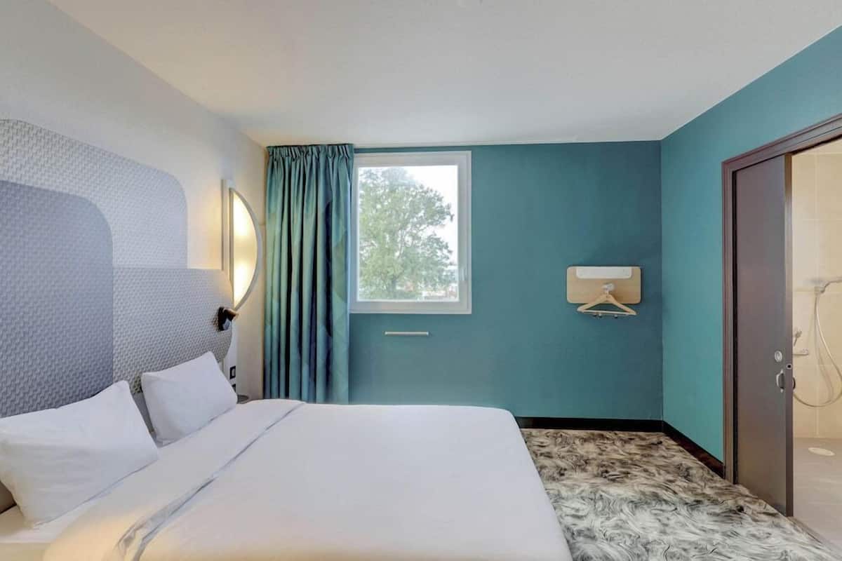Une chambre d’hôtel comprenant un lit, une fenêtre avec des rideaux, un luminaire fixé au mur et une porte donnant accès à une cabine de douche.
