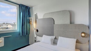 Une chambre d’hôtel avec un grand lit, une vue sur la ville par la fenêtre et une tête de lit moderne à motifs texturés.