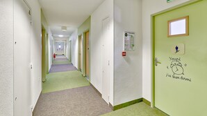 Un couloir d’hôtel avec de nombreuses portes, un détecteur d’incendie et une porte verte ornée d’une horloge et de l’inscription « Youpi, on va bien dormir ».