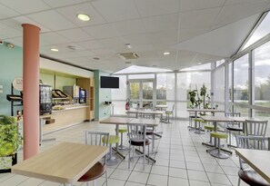 Een ruime binnenruimte met meerdere tafels en stoelen, grote ramen en een balie met een televisiescherm.