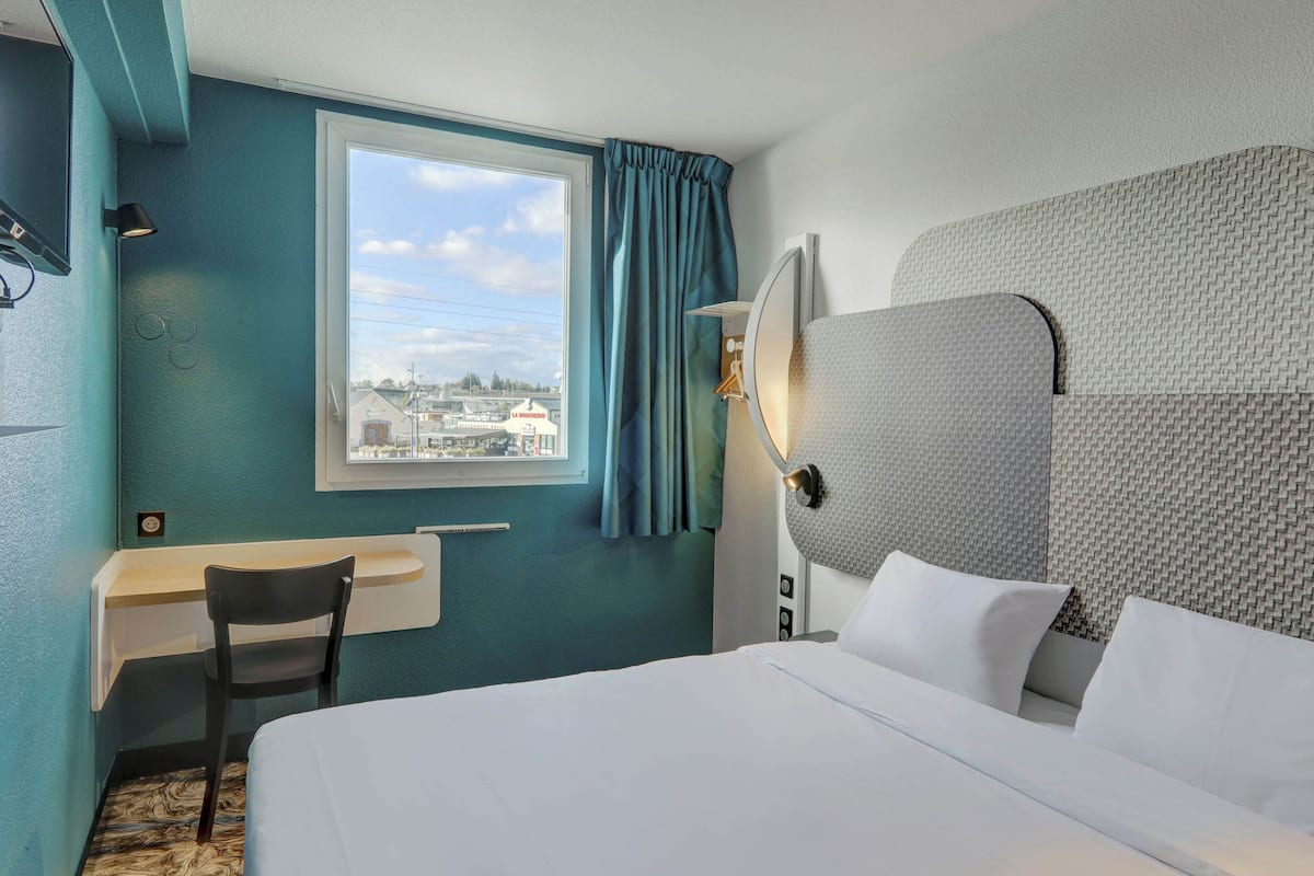 Habitación de hotel con cama, escritorio, silla, ventana con cortinas y vista panorámica de la ciudad.