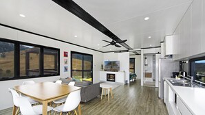 A modern kitchen with white cabinets, a dining area with a wooden table and white chairs, and a large window overlooking a scenic landscape. - Bindera Container House (BINDERA)