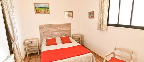 Un dormitorio con cabecera de madera, colcha roja, una silla de madera y una ventana con cortinas.