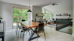 A spacious dining area with a wooden table and chairs, a ceiling fan, and large windows offering a view of greenery.