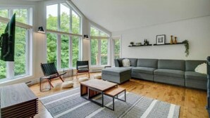 A spacious living room with large windows, a grey sofa, a wooden coffee table, and a black chair.