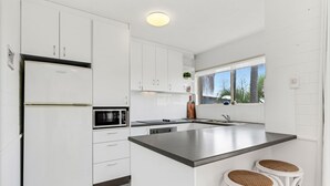 A modern kitchen with white cabinetry, a stainless steel island, and a window with a view of palm trees. - Coolum Beach 1 Bedroom Apartment Ocean View @ Surf Dance (Sunshine Coast)