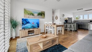 A modern living room with a TV, wooden furniture, and a kitchen in the background. - Coolum Beach 1 Bedroom Apartment Ocean View @ Surf Dance (Sunshine Coast)