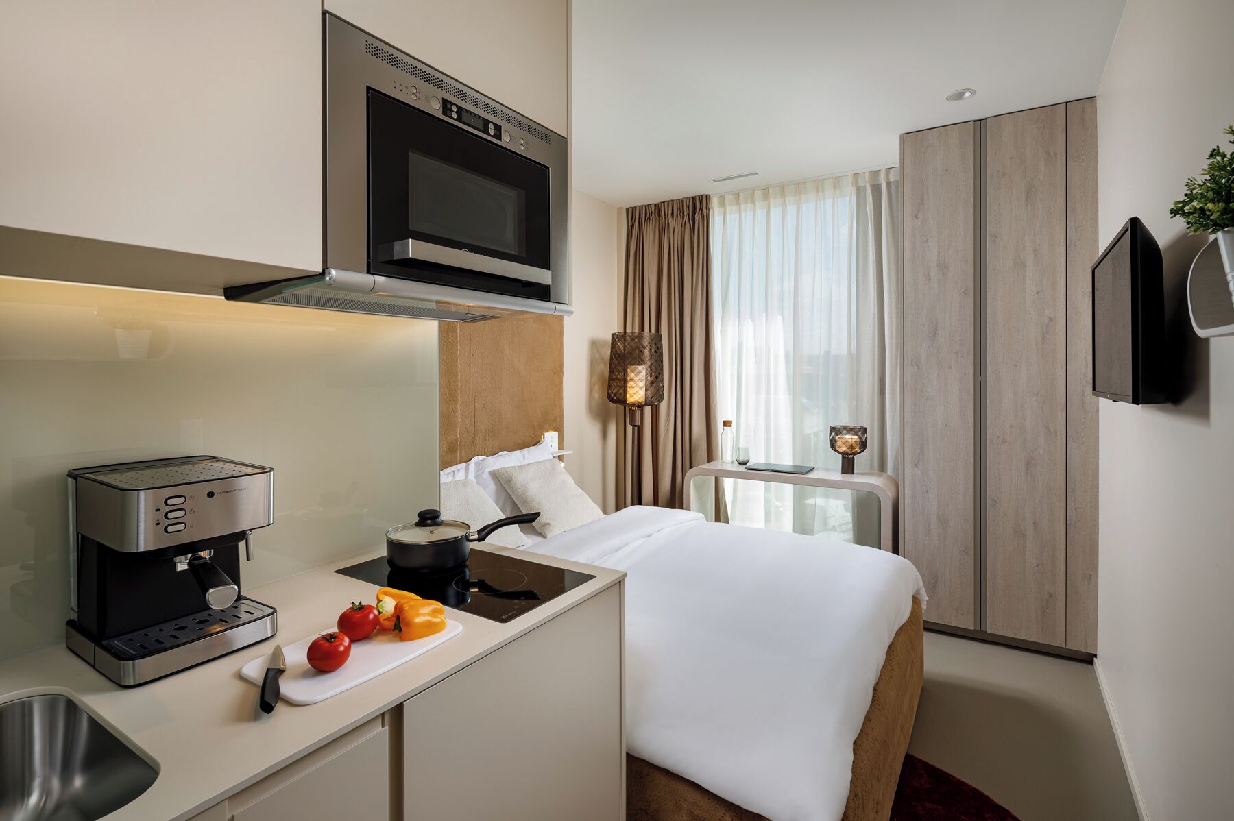 Una habitación de hotel compacta con kitchenette, una cama, una mesita de noche, un televisor y una ventana con cortinas.