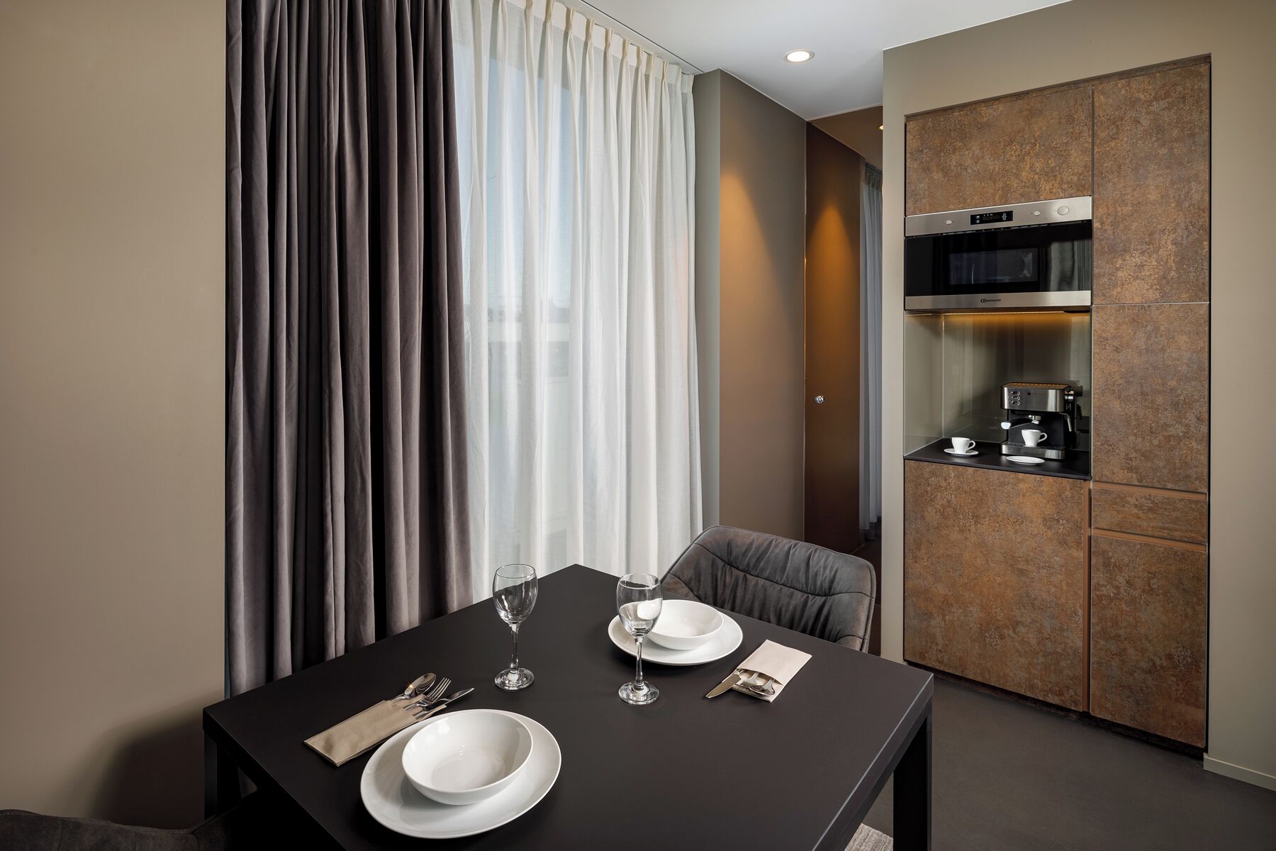 A modern hotel room with a dining table set for two, a coffee machine, and a built-in oven.