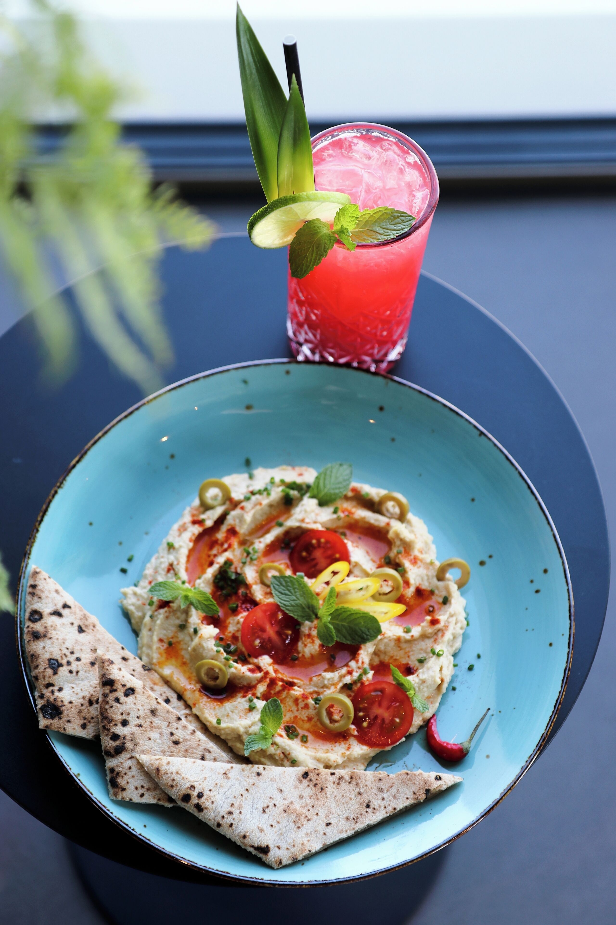 A plate of hummus with tomatoes, olives, and mint, accompanied by flatbread, and a glass of red drink with a mint garnish.