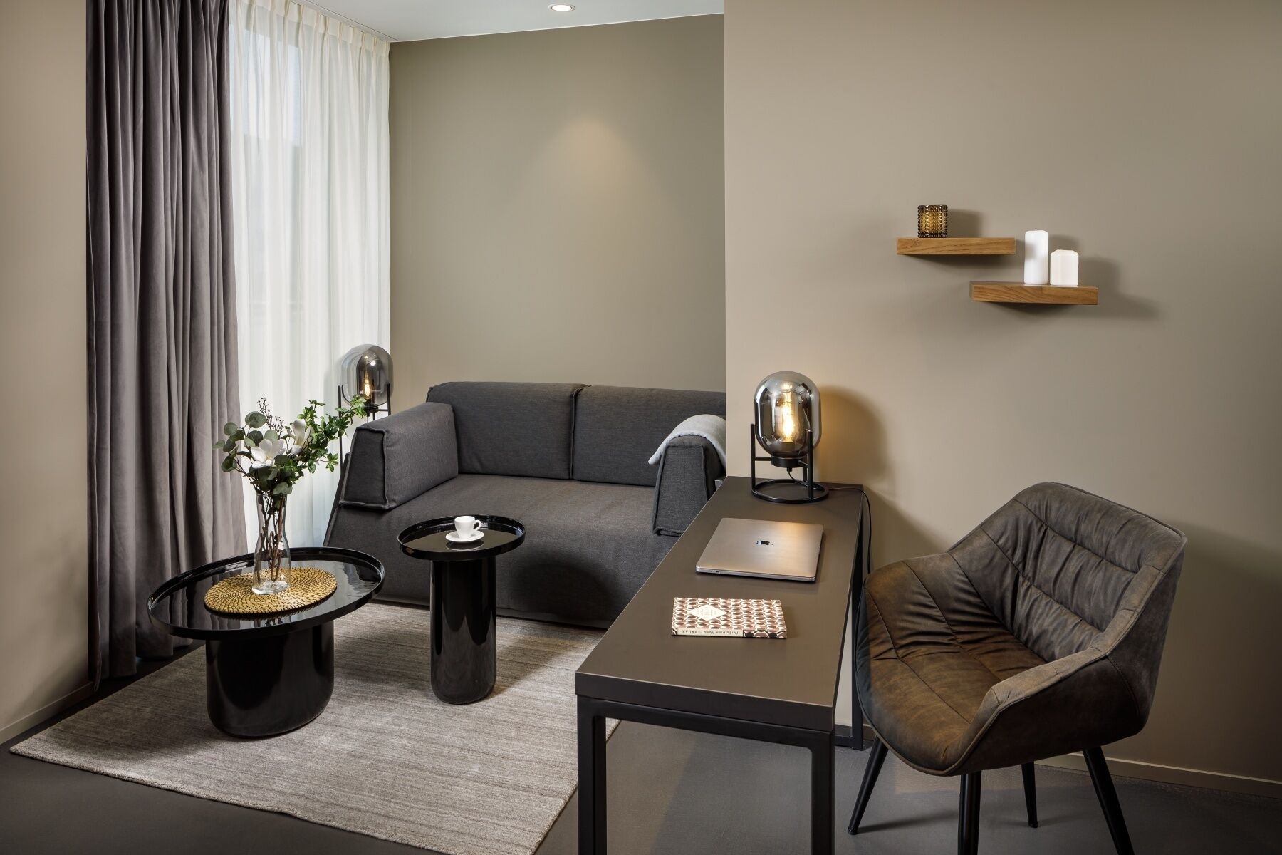 A modern living room with a grey sofa, a round black coffee table, a laptop on a rectangular table, a black chair, and a decorative lamp.