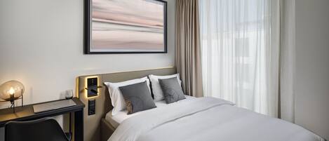A hotel room with a bed, a desk, a chair, a framed picture on the wall, and a window with curtains.