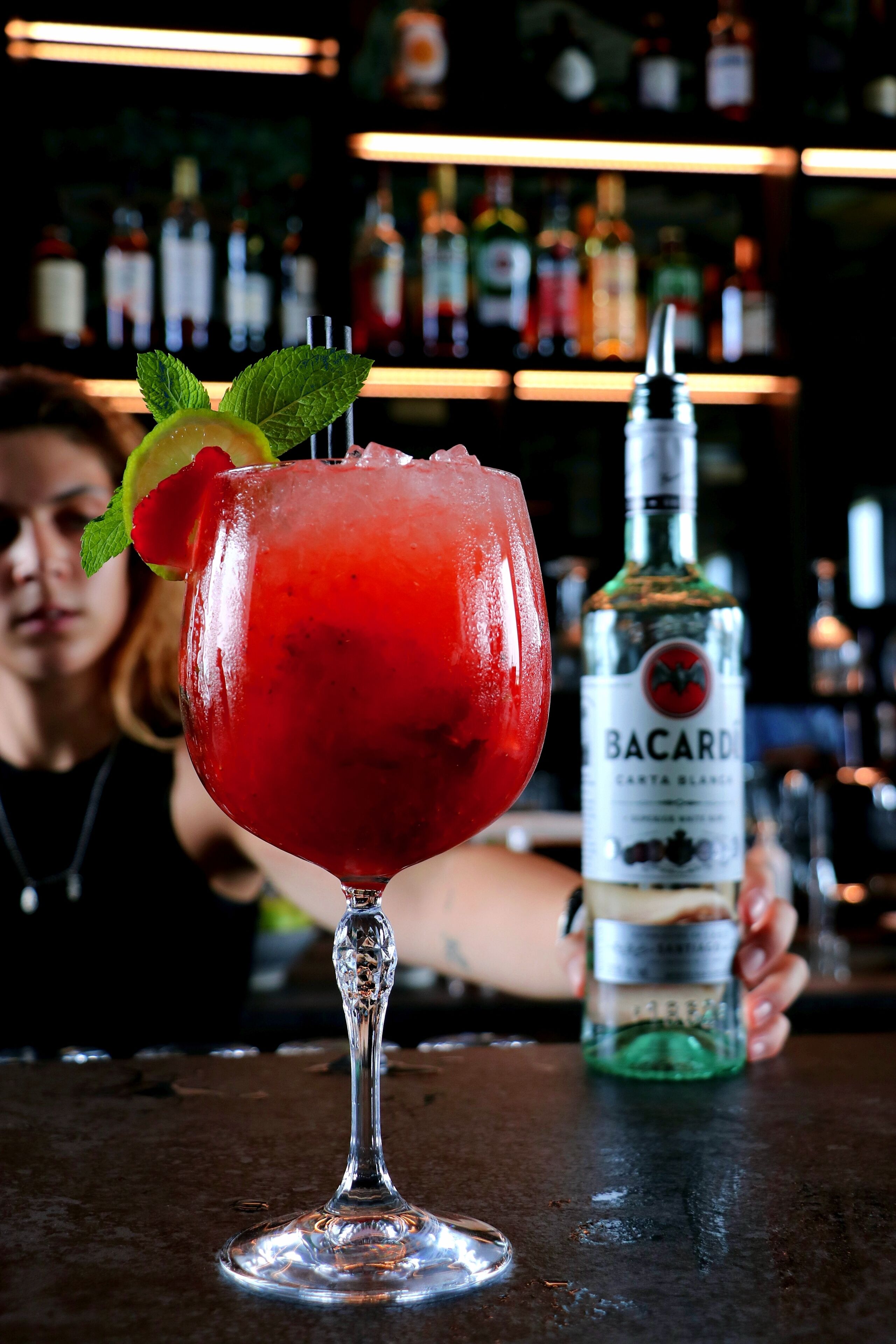 Un cocktail servi dans un verre à pied, décoré d’une feuille de menthe, une bouteille de Bacardi en arrière-plan et une personne floue derrière le bar.